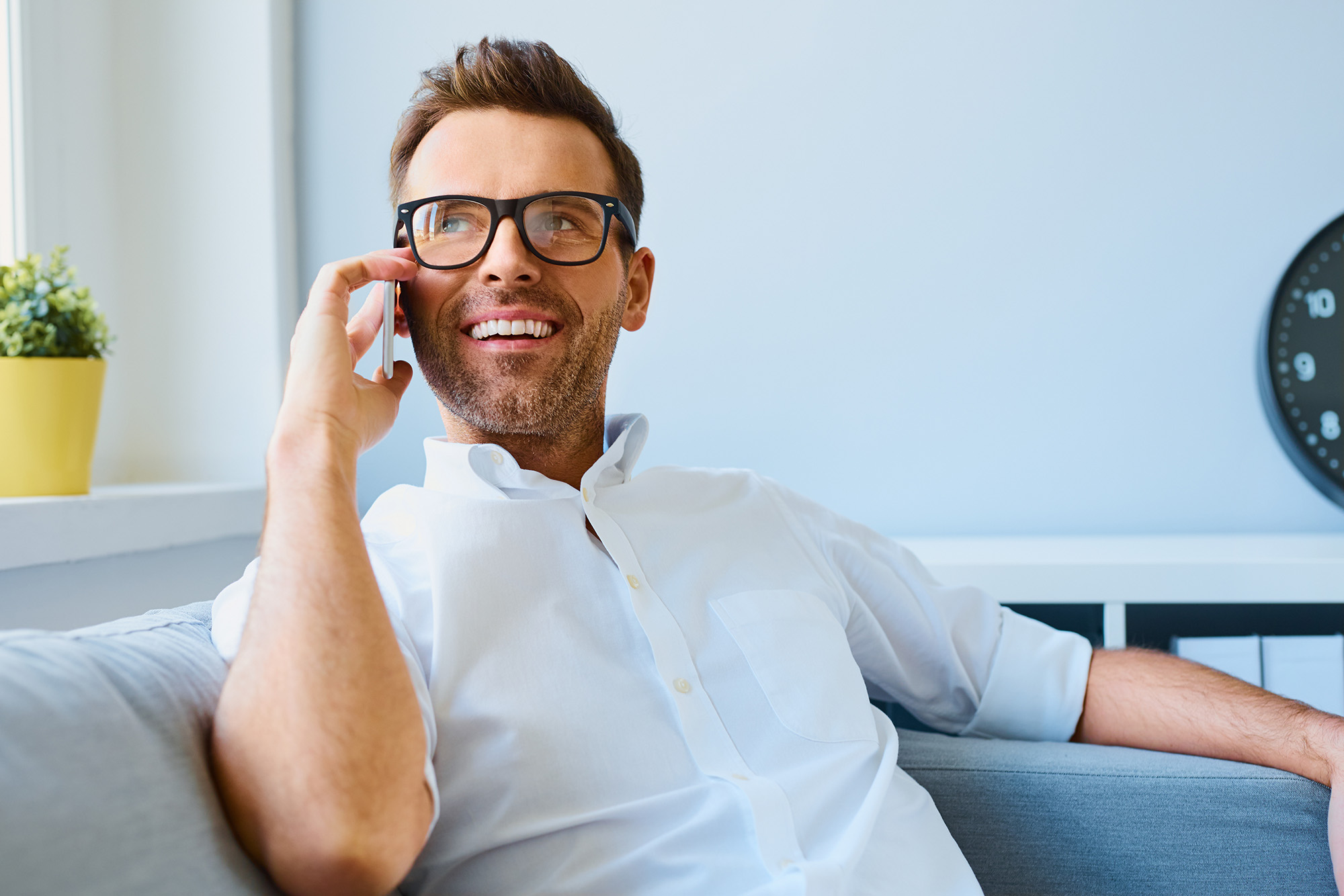 Młody szczęśliwy mężczyzna w okularach siedzi zrelaksowany na sofie i rozmawia przez telefon